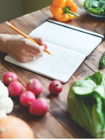 writing in a notepad on a wooden chopping board with vegetables around it